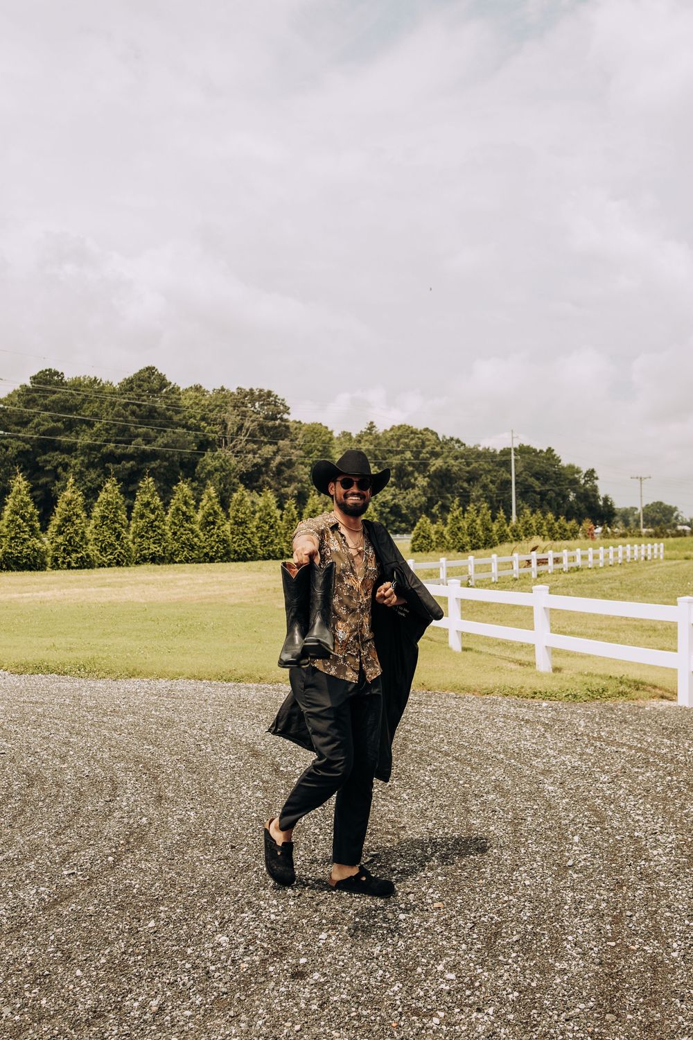 Stylish man in a cowboy hat and patterned shirt walking along a gravel path by a white fence.