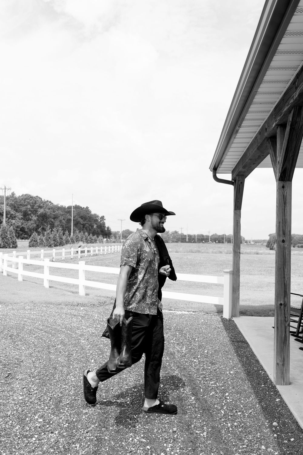 Black and white photo of a man in a cowboy hat walking near a white fence outdoors.