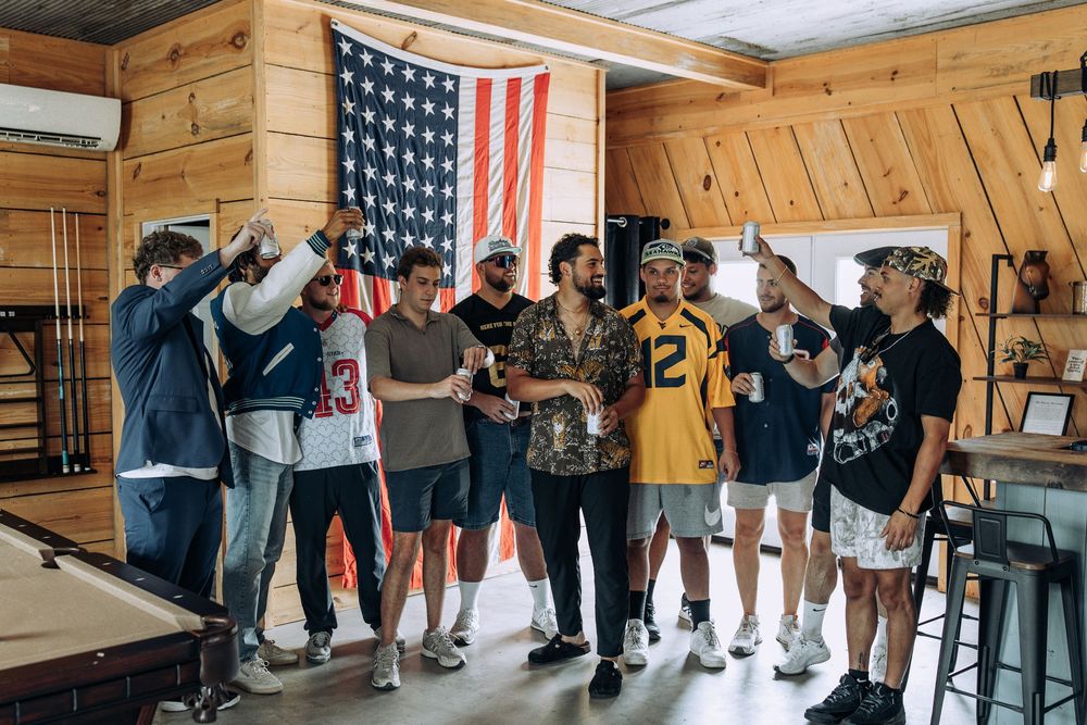 Group of groomsmen in casual attire raising drinks in a rustic wood-paneled room with American flag.