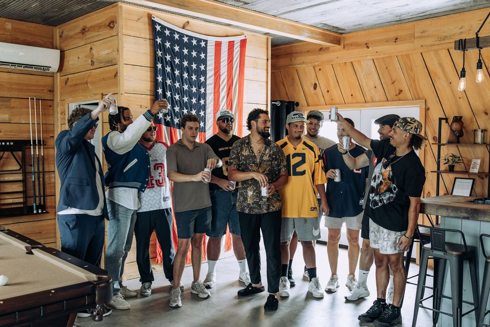 Groomsmen in sports jerseys toasting together in a wood-paneled room with an American flag backdrop.