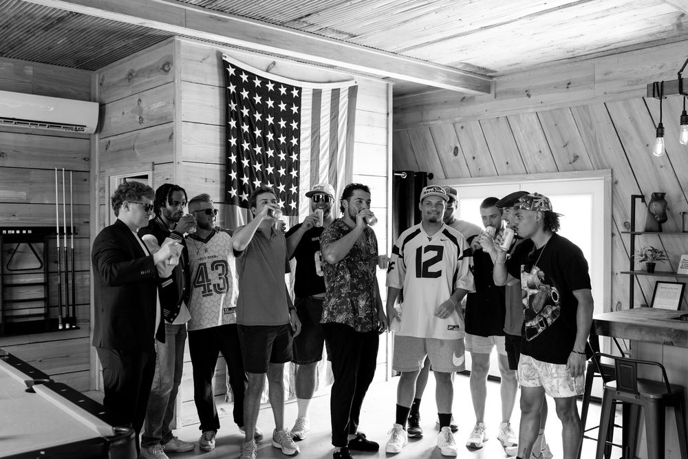Black and white photo of groomsmen toasting drinks indoors with American flag backdrop at rustic cabin wedding.