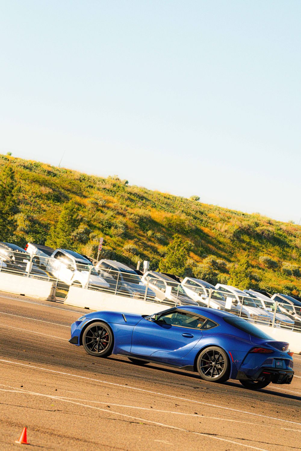 A sleek blue BMW sports car driving on a dirt track with hills and parked cars in the distance.