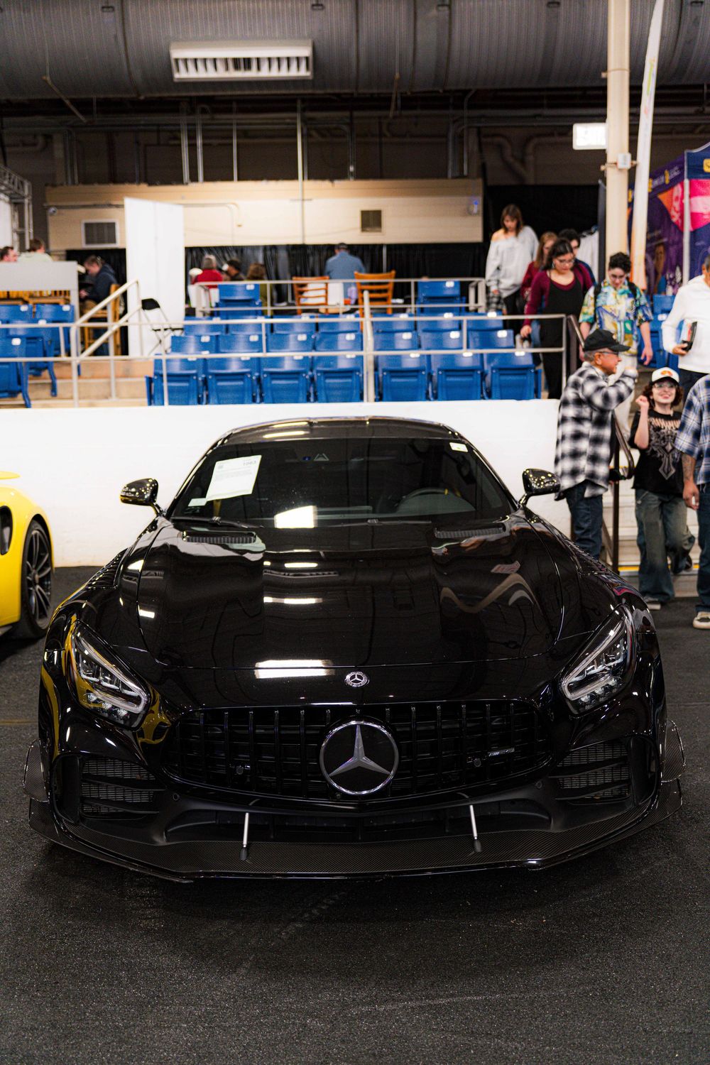 Sleek black Mercedes-AMG GT sports car on display at an indoor auto show with blue stadium seating in background.