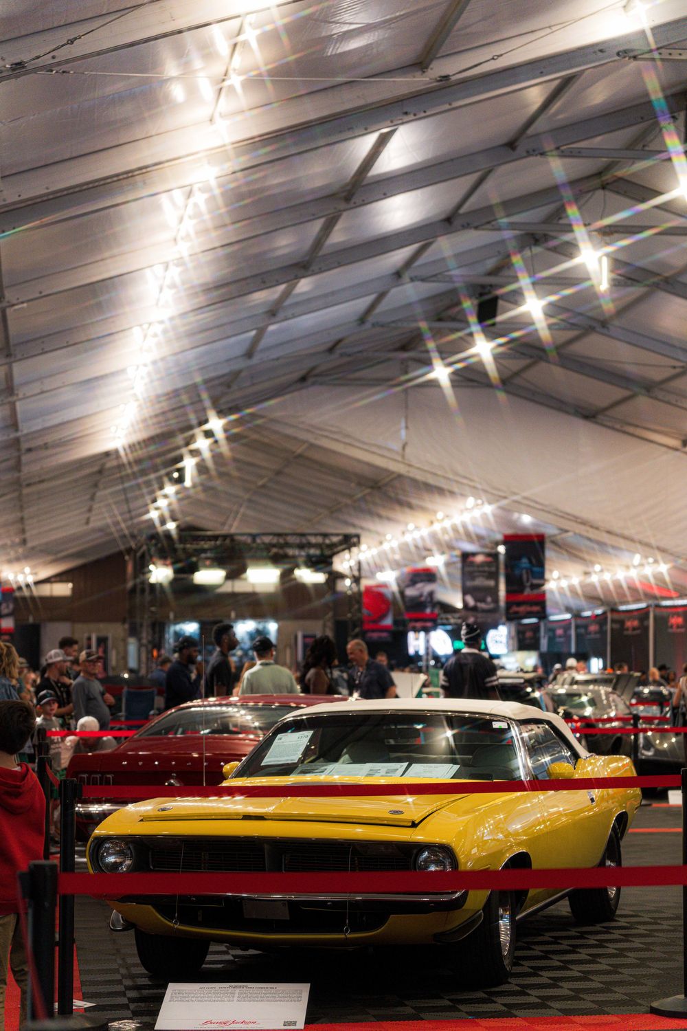 Classic muscle cars on display in a large indoor exhibition tent with overhead lighting.