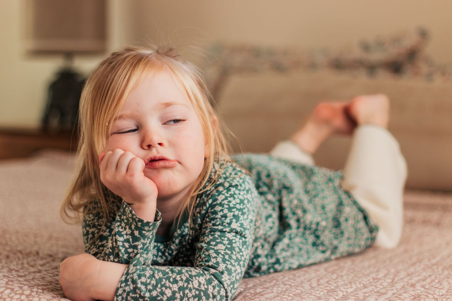 Bored little blonde girl lying on a bed resting her chin on her hand, wearing a floral pajama top | Kids Birthday Photo Sessions