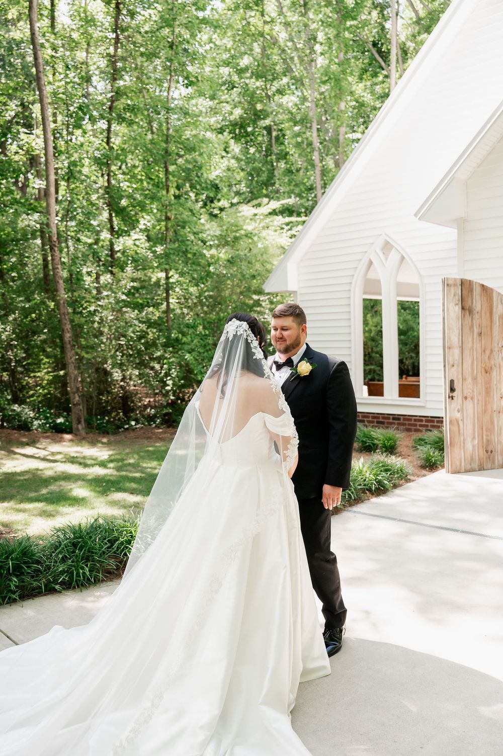 A wedding first look sequence outside a white chapel building surrounded by trees.