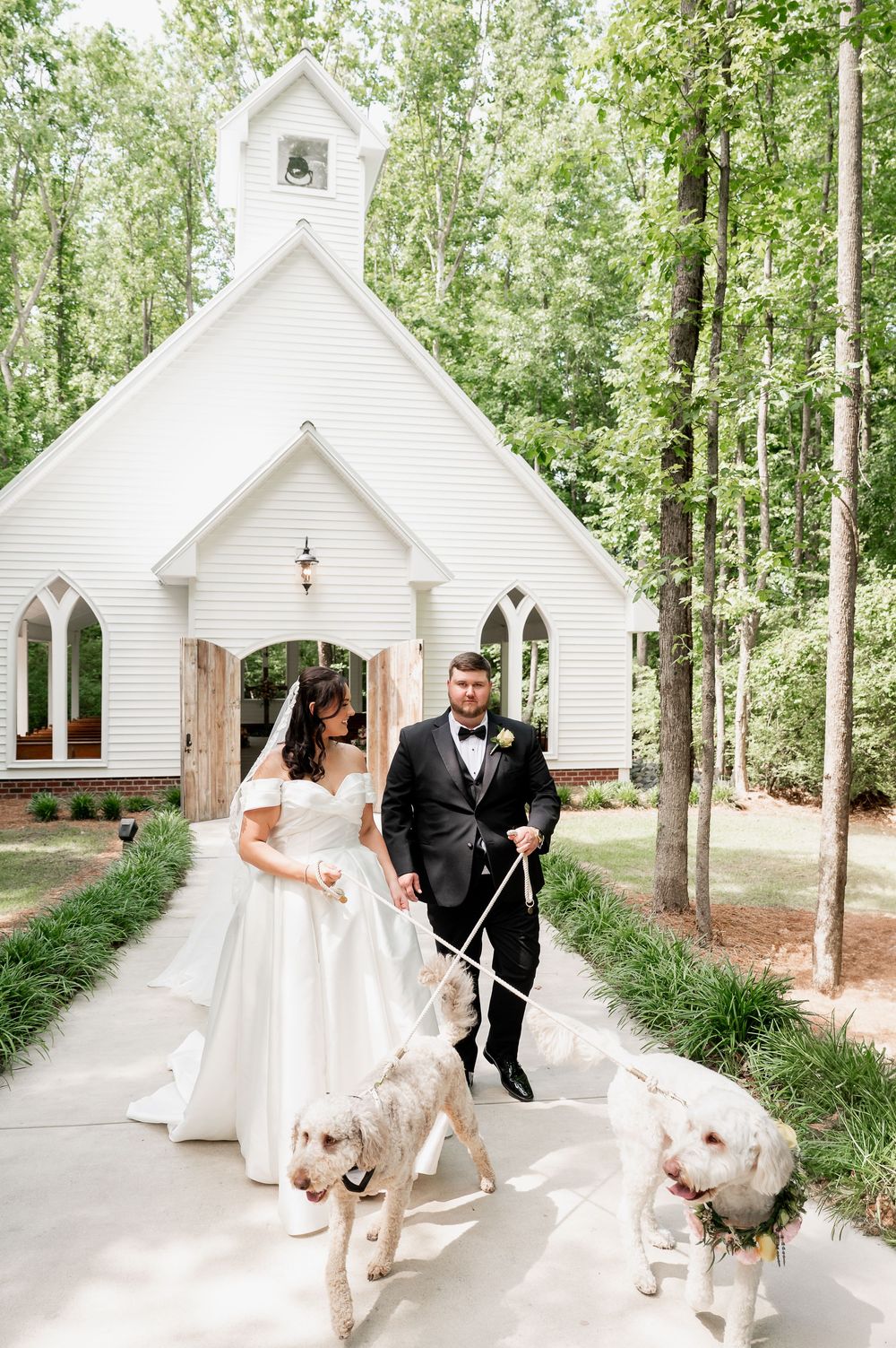 A couple in wedding attire walks with their two white dogs outside a charming white chapel in a wooded setting.