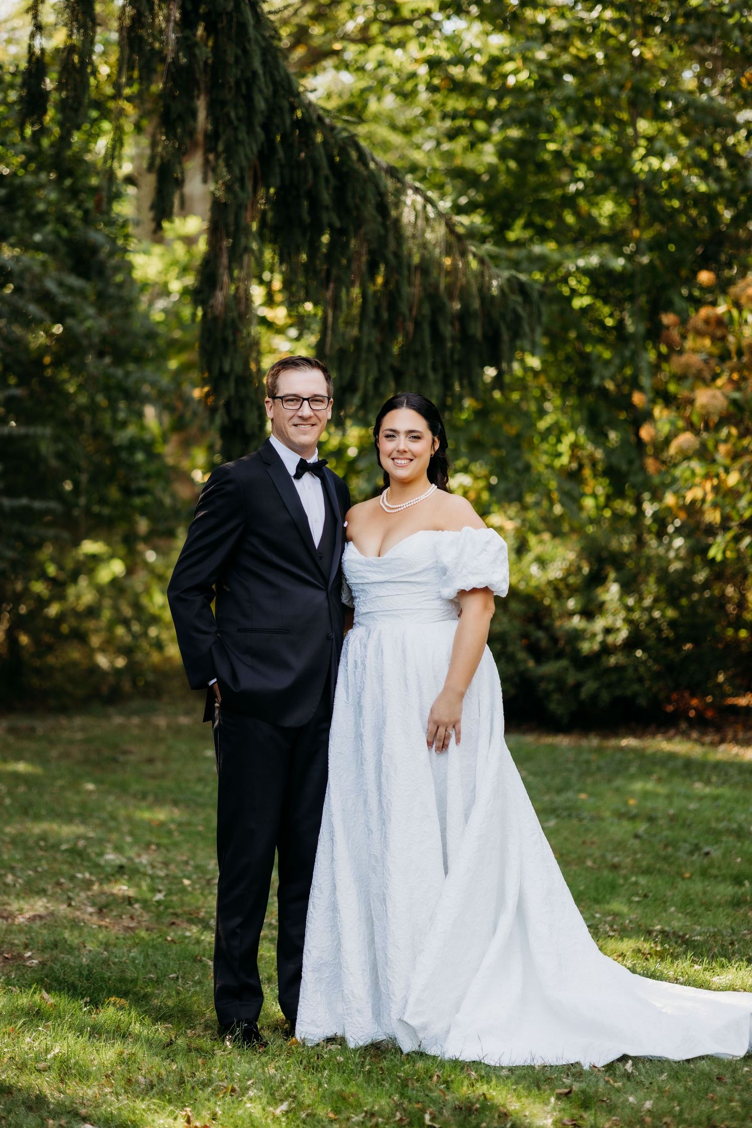 A couple in wedding attire poses together outdoors in a wooded setting with sunlit trees in the background.