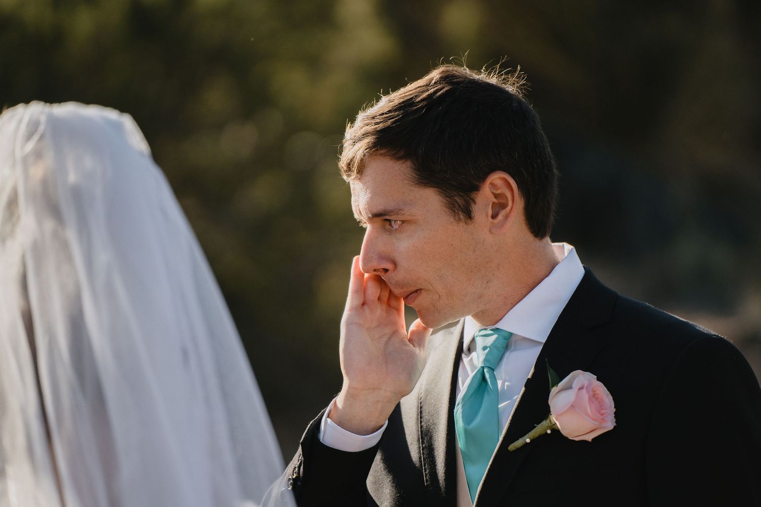 Emotional groom wiping tears during outdoor wedding ceremony wearing black suit with mint tie and pink boutonniere.