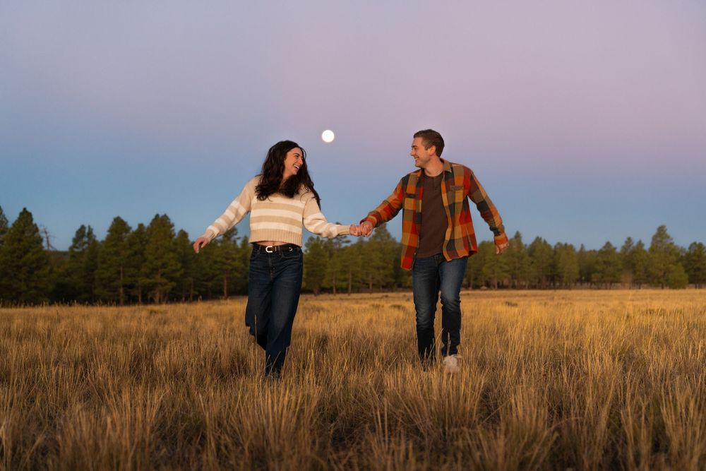 Couple holding hands while walking through golden grass field at sunset.