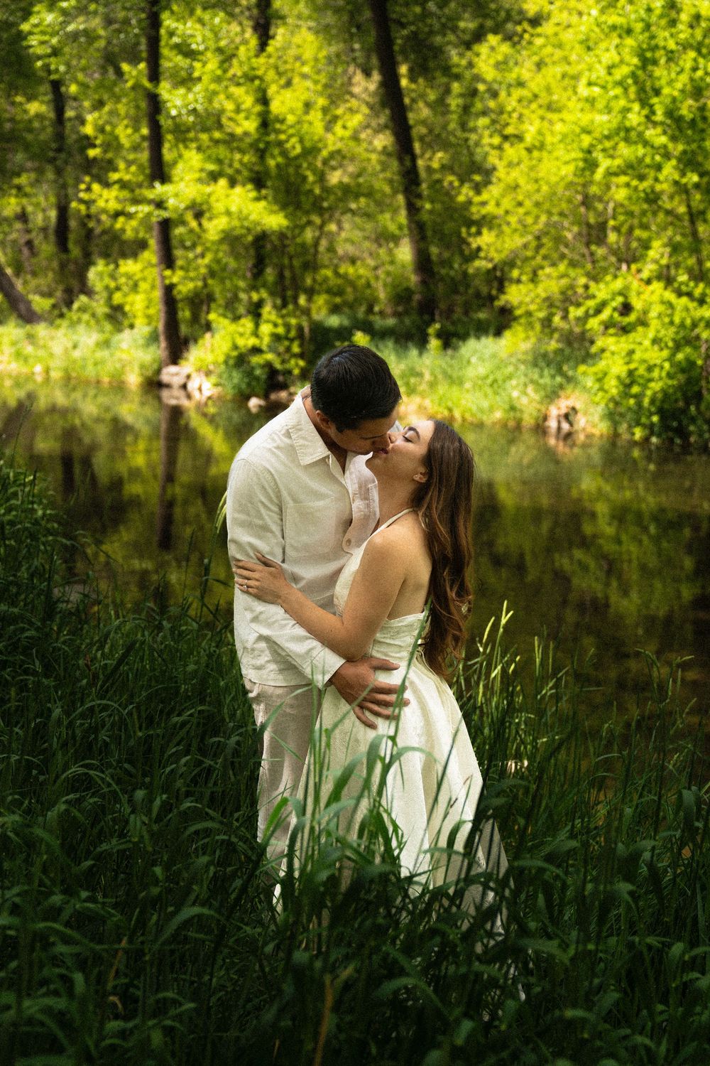 Romantic couple embracing in tall grass by sunlit pond.