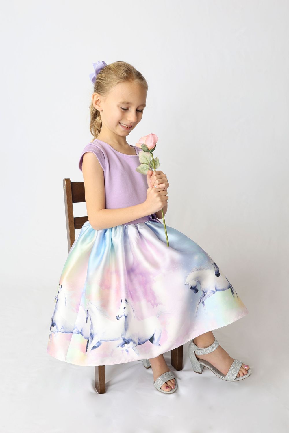 Young girl in pastel dress with watercolor pattern sits on wooden chair holding pink flower against white background.