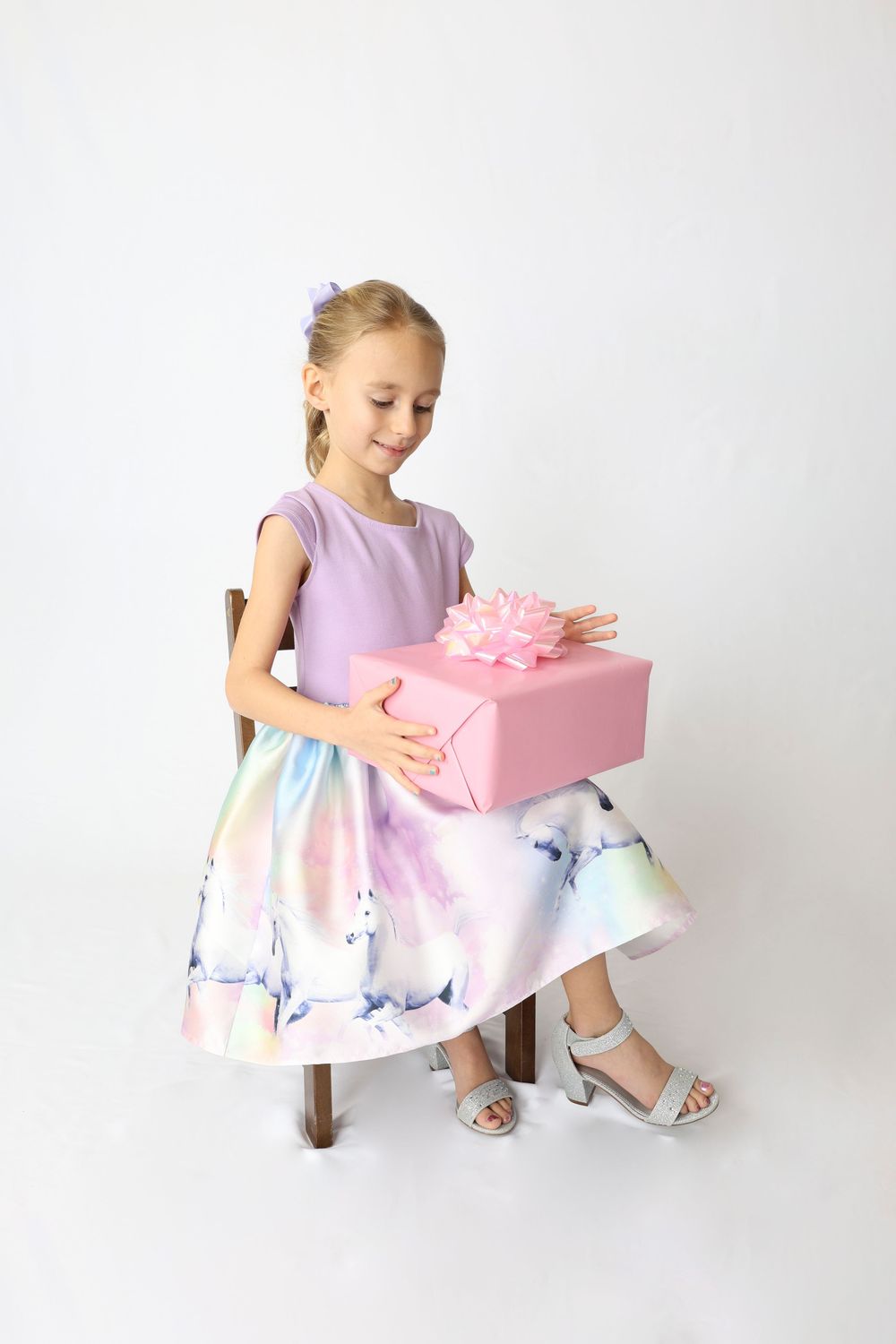 A small child in a purple and white dress sits with a pink gift box in a bright studio setting.