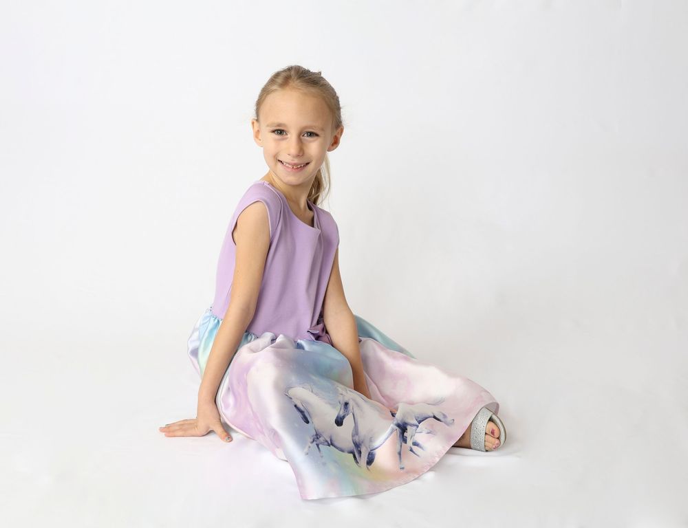 A child in a lavender dance dress sits gracefully on the floor in a bright studio setting.