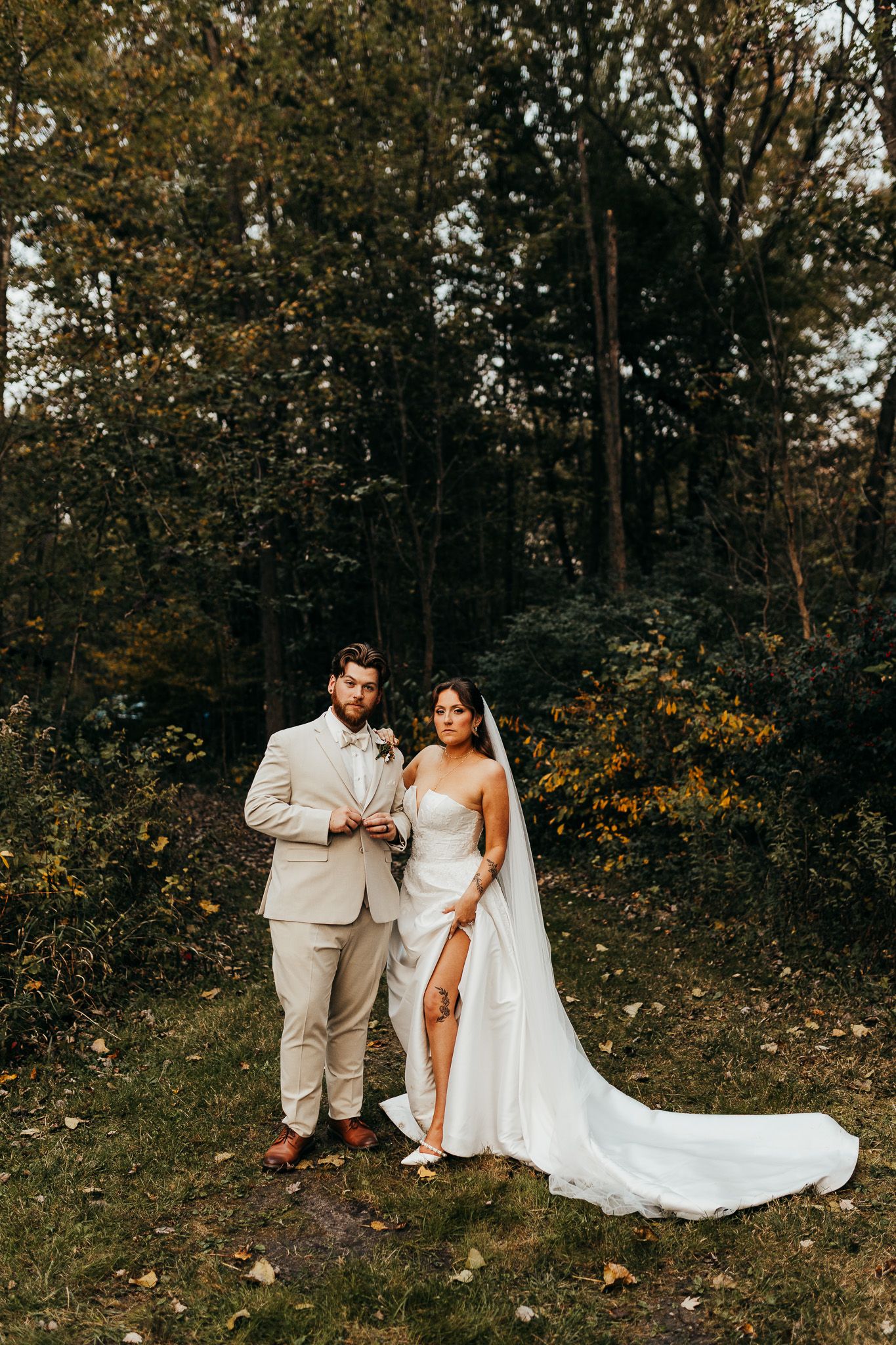 A couple in wedding attire poses together in a wooded outdoor setting with fall foliage in the background.