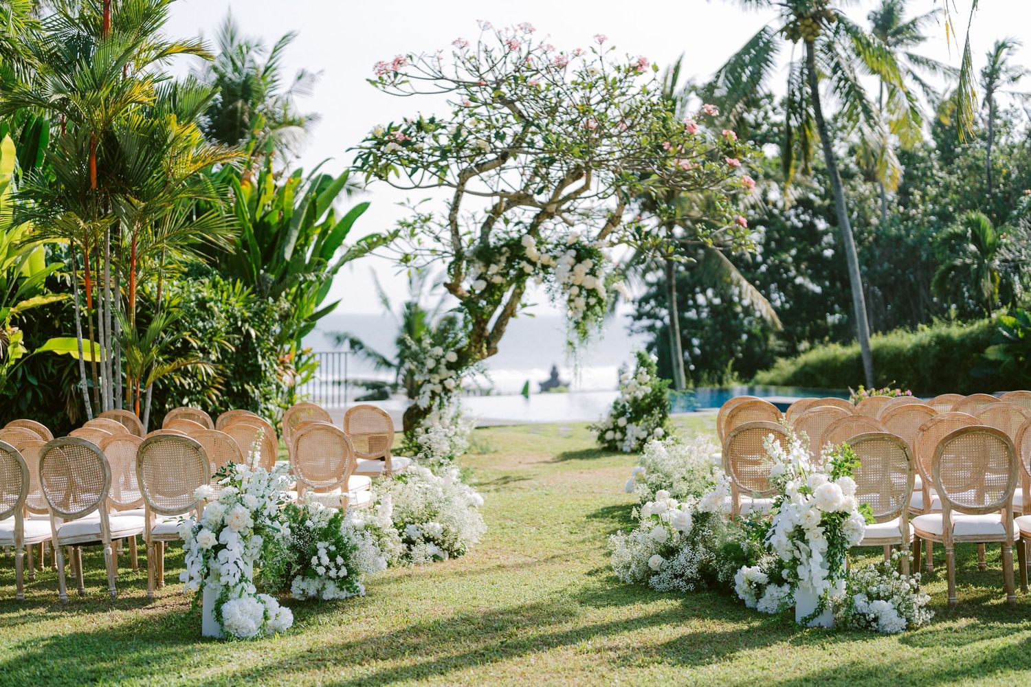 Elegant outdoor wedding ceremony setup with rattan chairs and white floral arrangements on a tropical beachfront lawn.