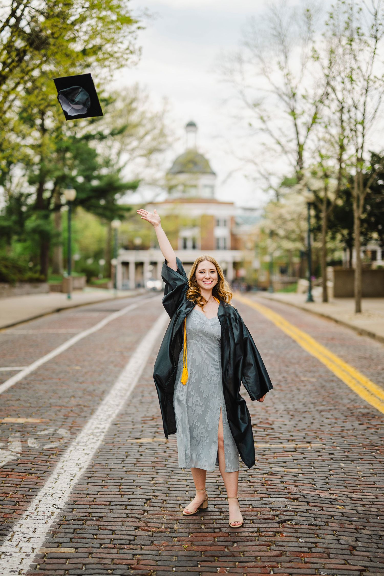 Graduate in a gray dress and black graduation gown celebrates on a brick-paved walkway with historic buildings in background.