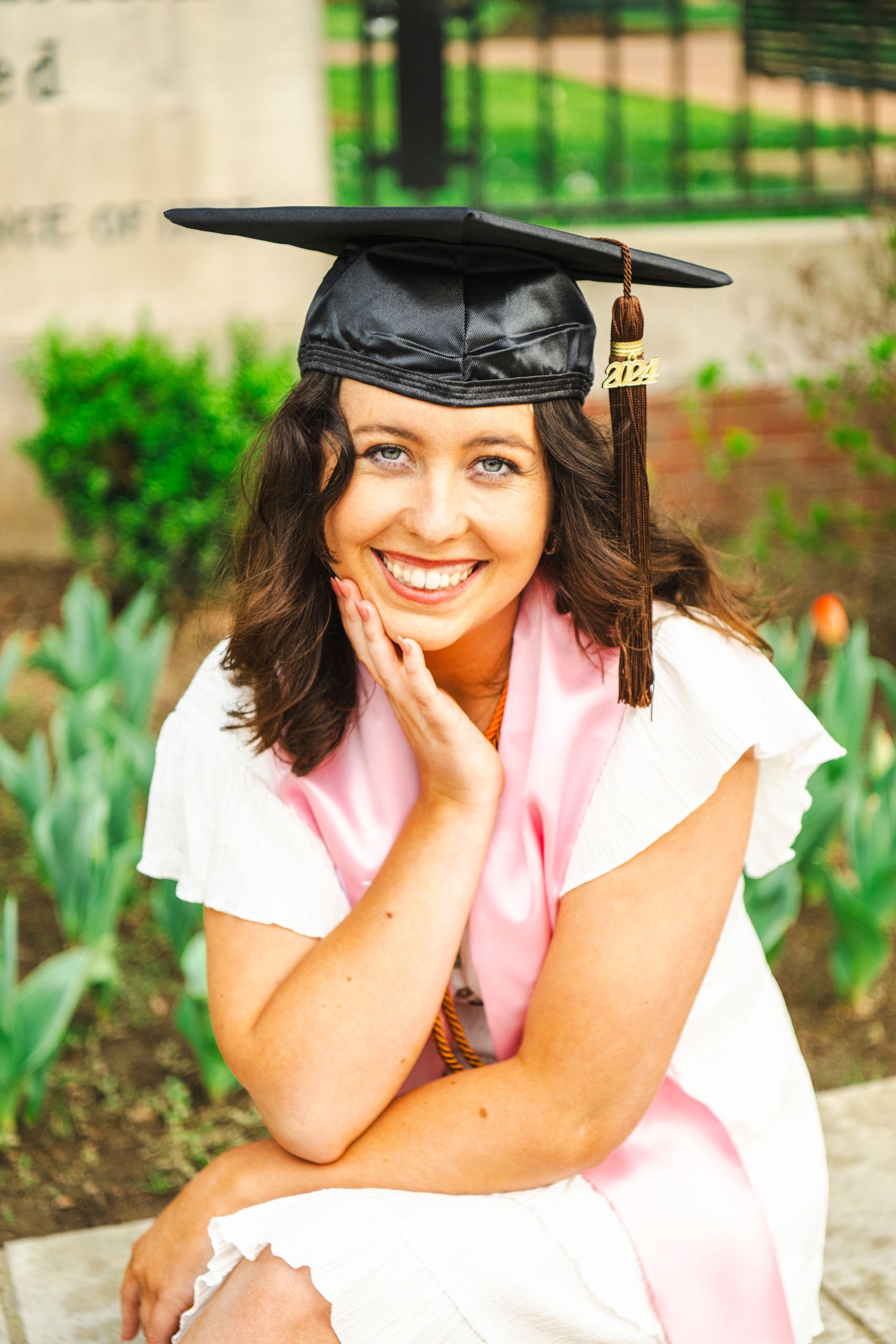 Graduate in white dress and black cap poses on ground with pink sash for outdoor portrait session.