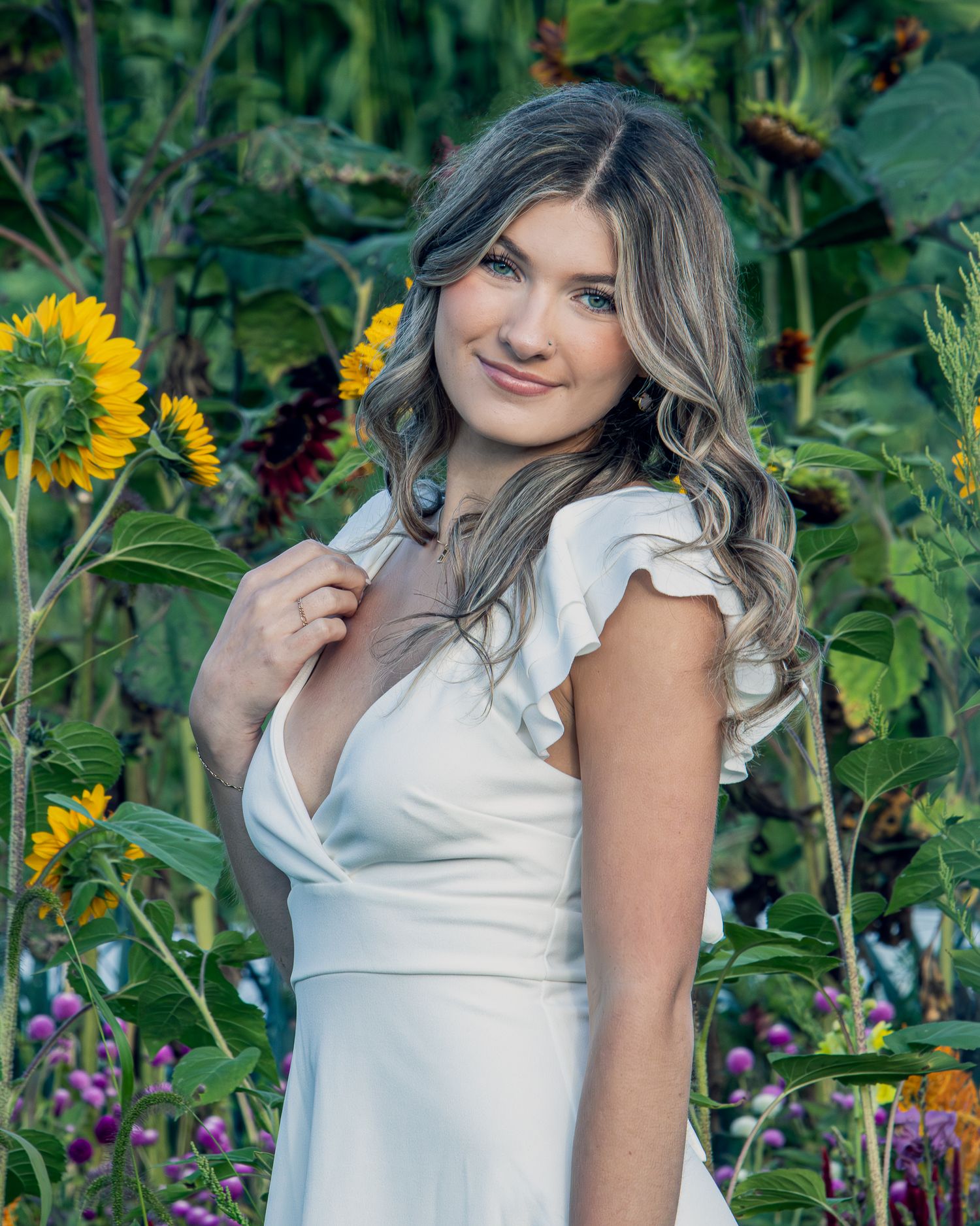 A woman in a white dress poses among vibrant sunflowers and purple flowers in a garden setting.