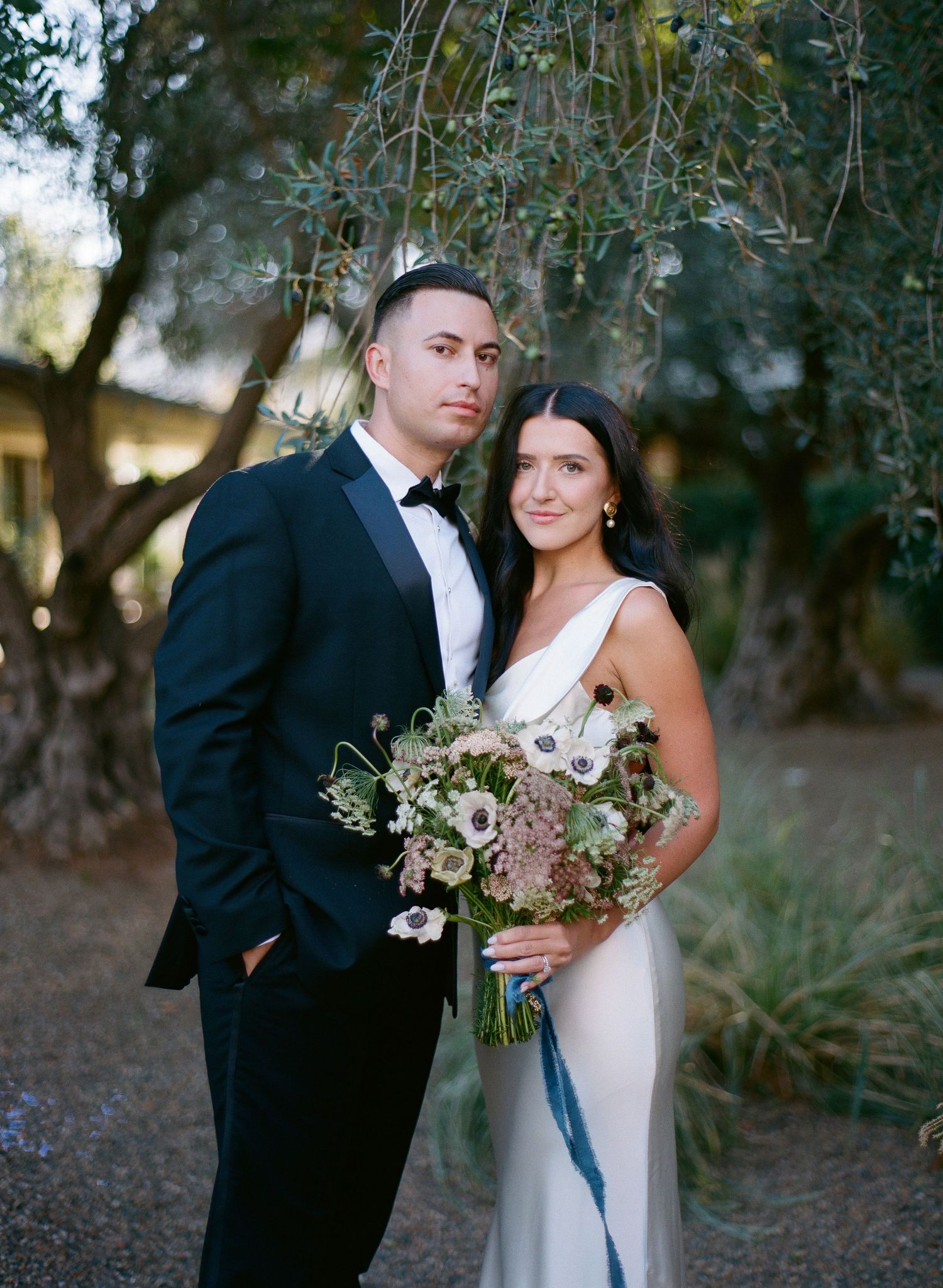 A couple stands outdoors beneath hanging trees with a natural floral bouquet.