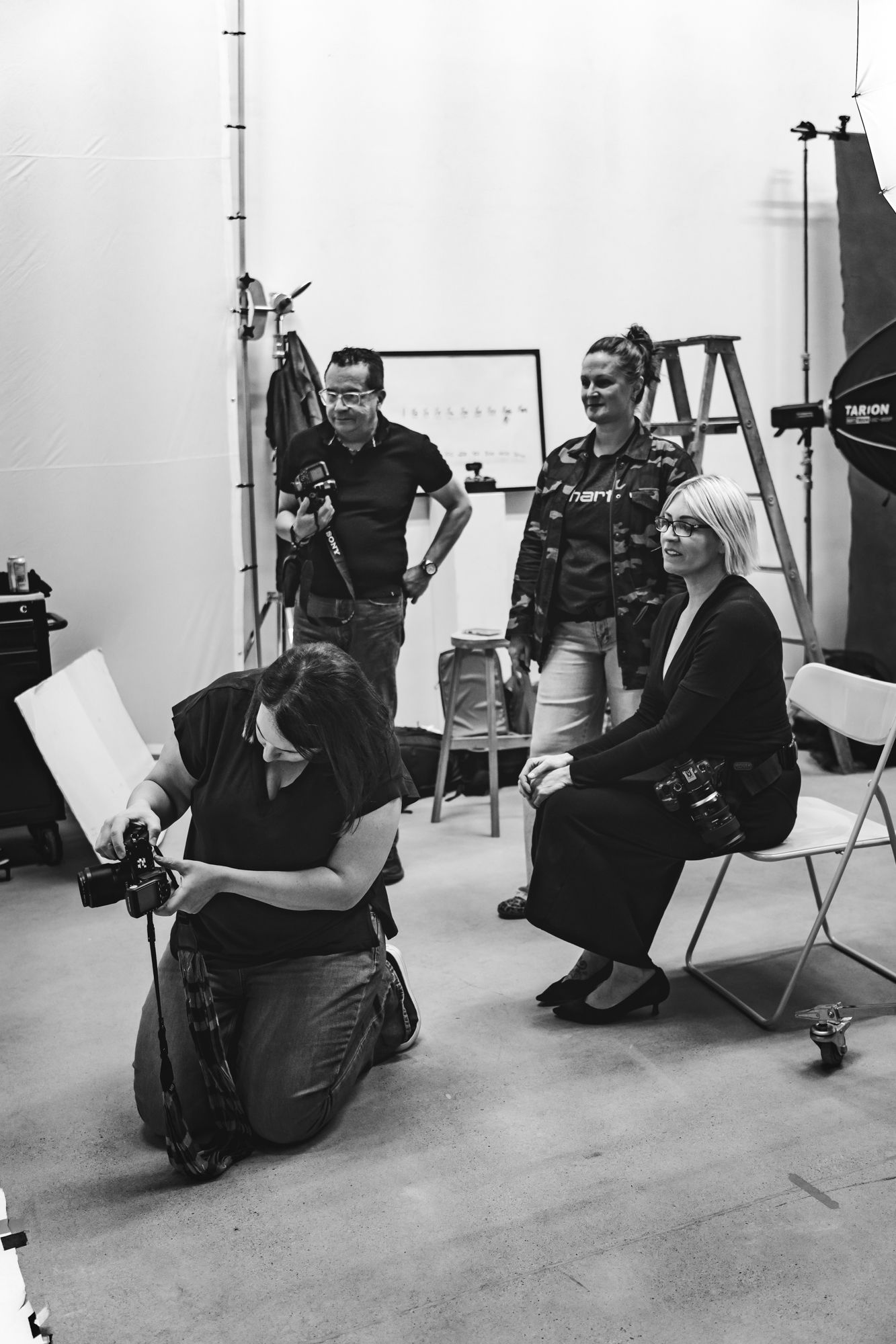 Professional photographer in black attire crouches and poses while taking photos in a white studio setting.