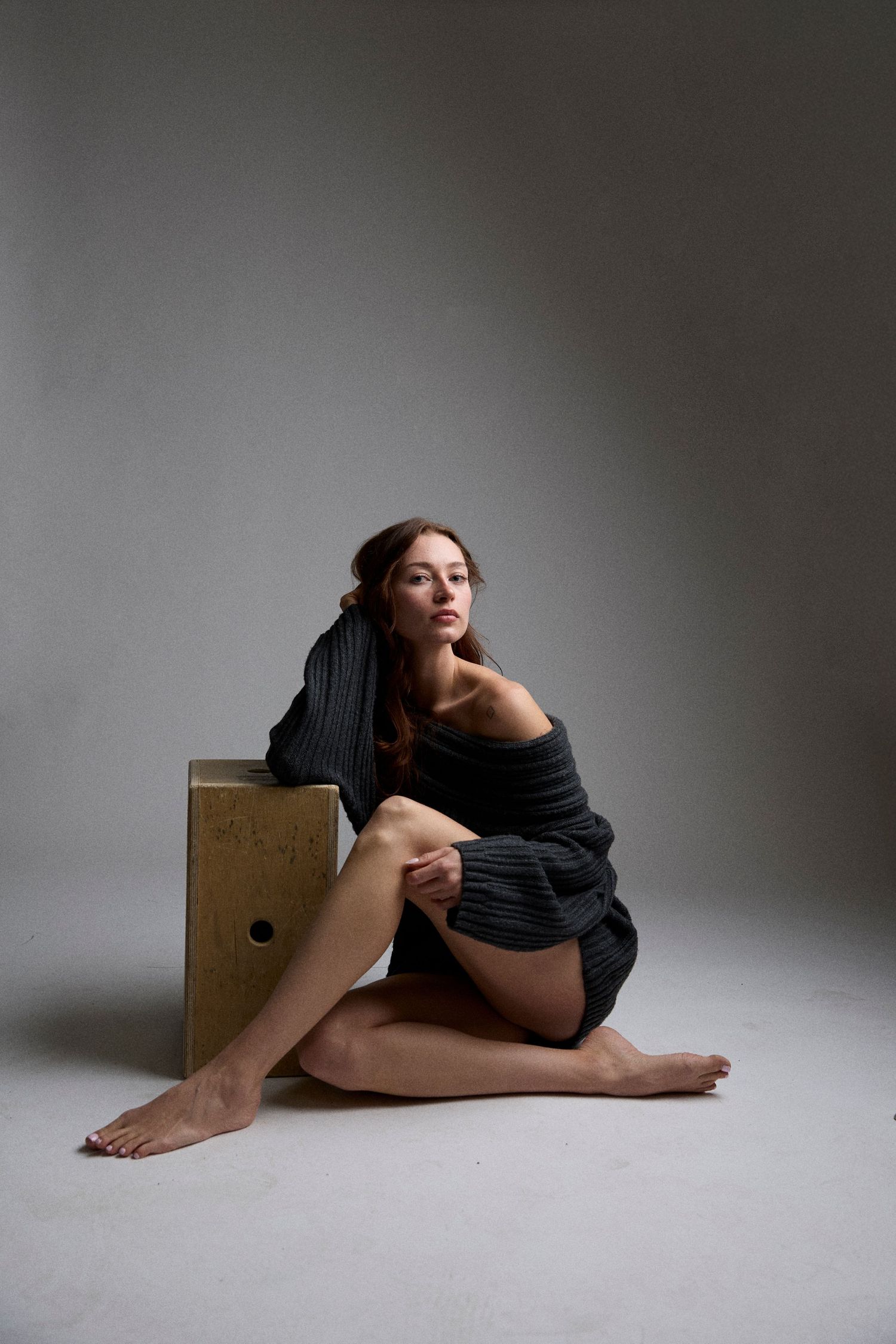 A person in black clothing lies on a light floor against a gray background in a minimalist artistic pose.