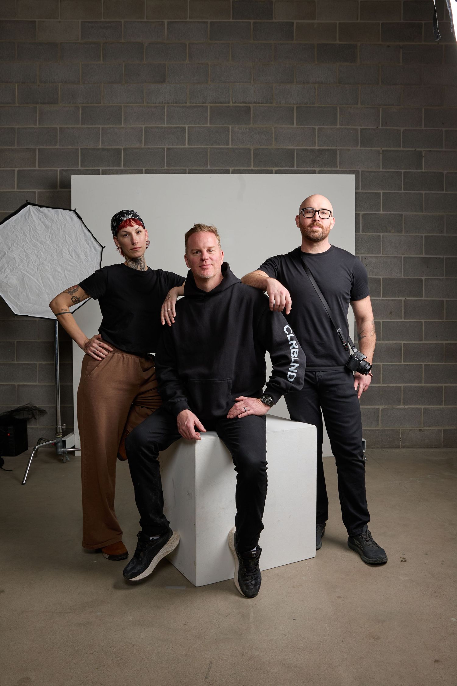 Three people in black clothing pose together on a white cube in an industrial space with concrete walls and floor.
