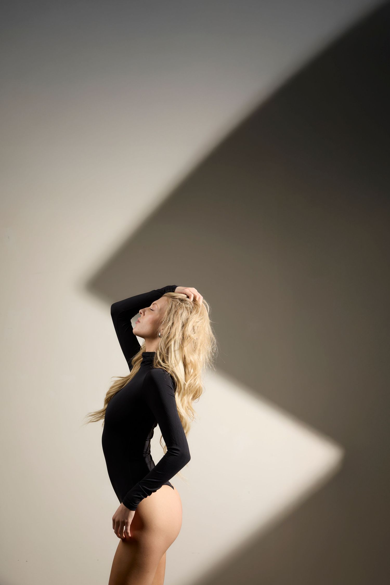 A figure in a black bodysuit poses dramatically against a white wall with angular shadows creating artistic contrast.