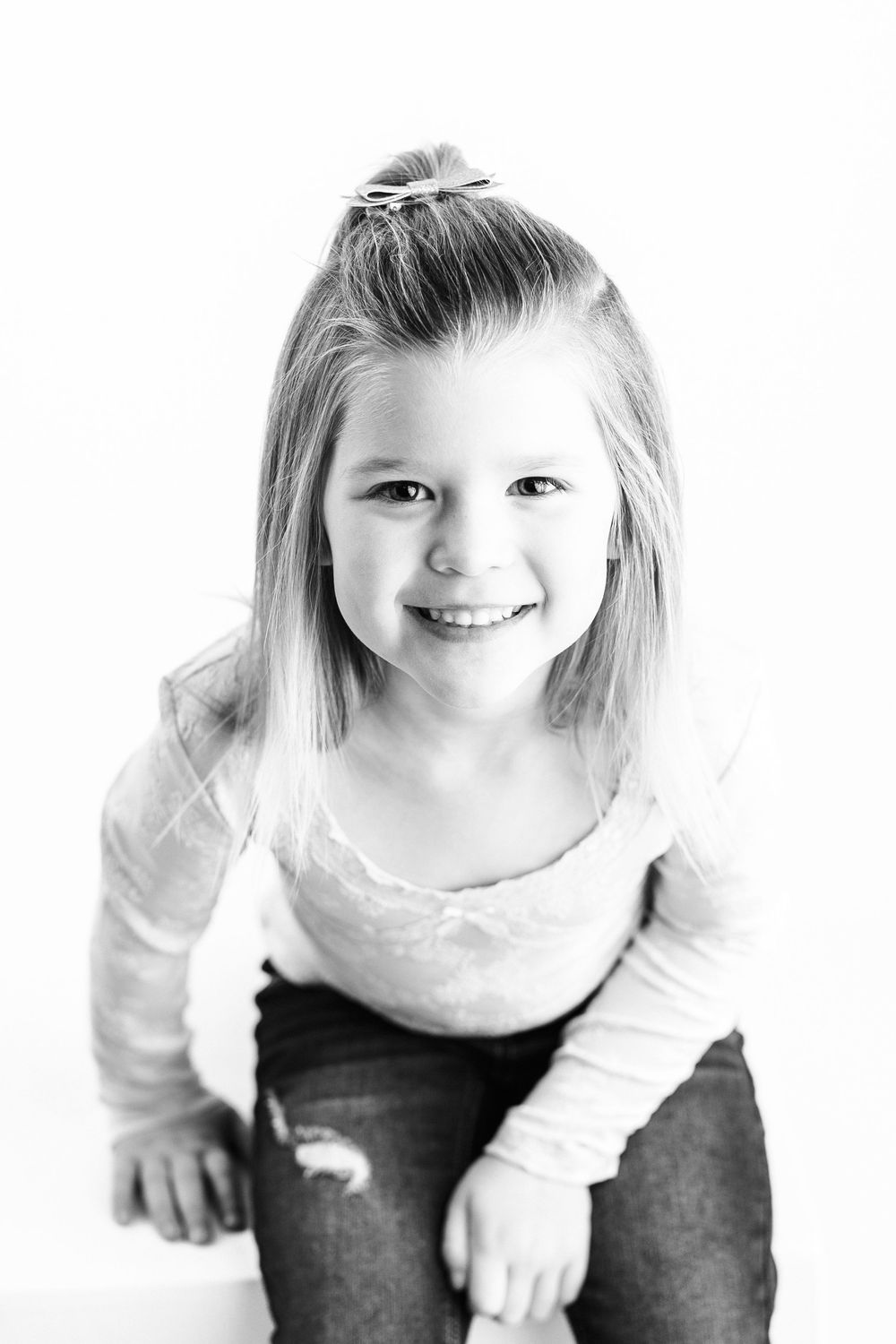 Black and white portrait of a child in a white shirt sitting against a light background, smiling joyfully.