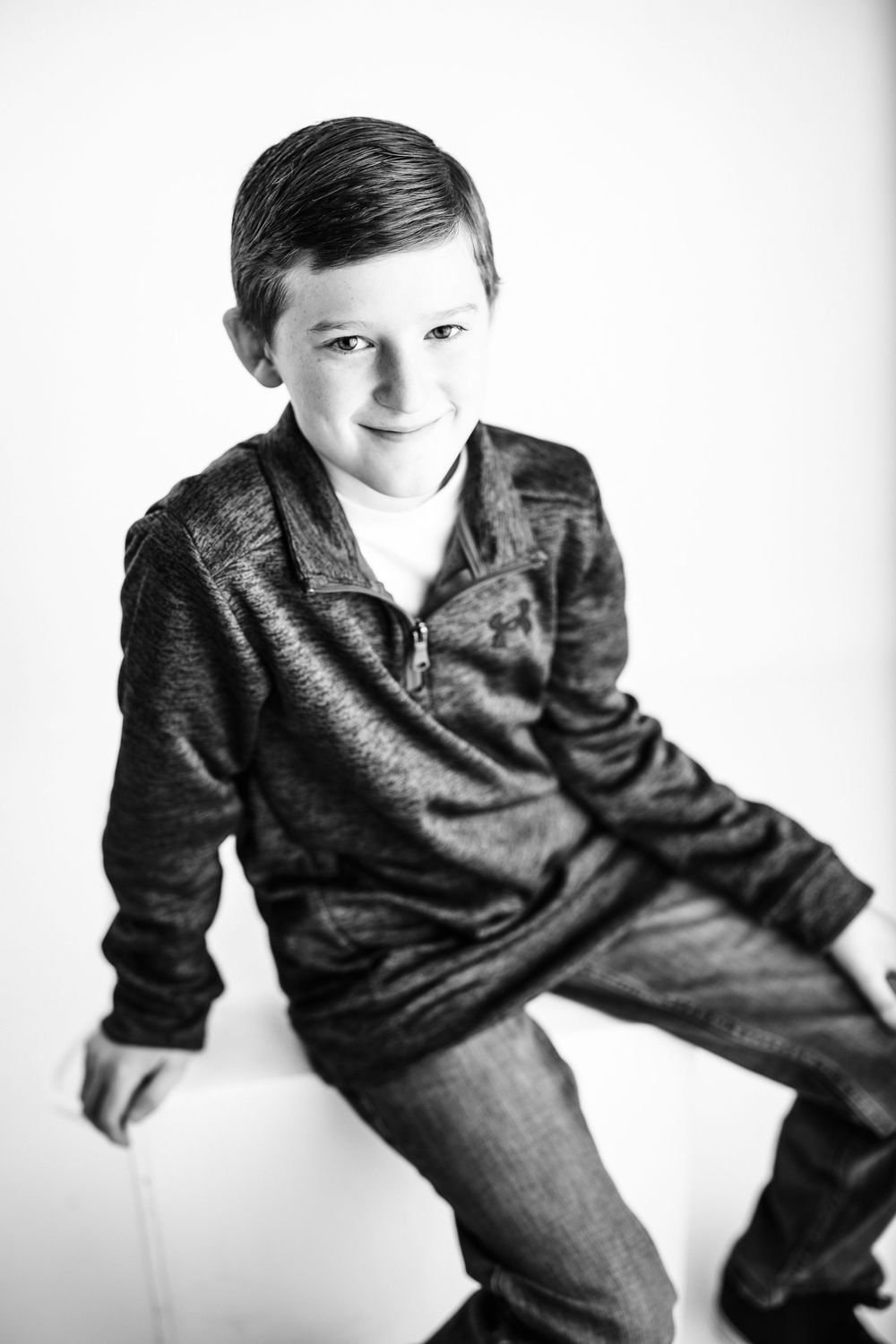 Artistic black and white studio photo of a child in casual clothing posing on a white backdrop.