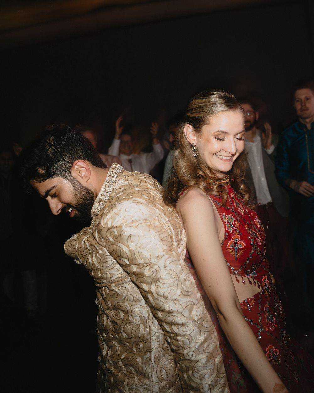 A joyful bride in red and groom in gold sherwani share a fun back-to-back dance moment.