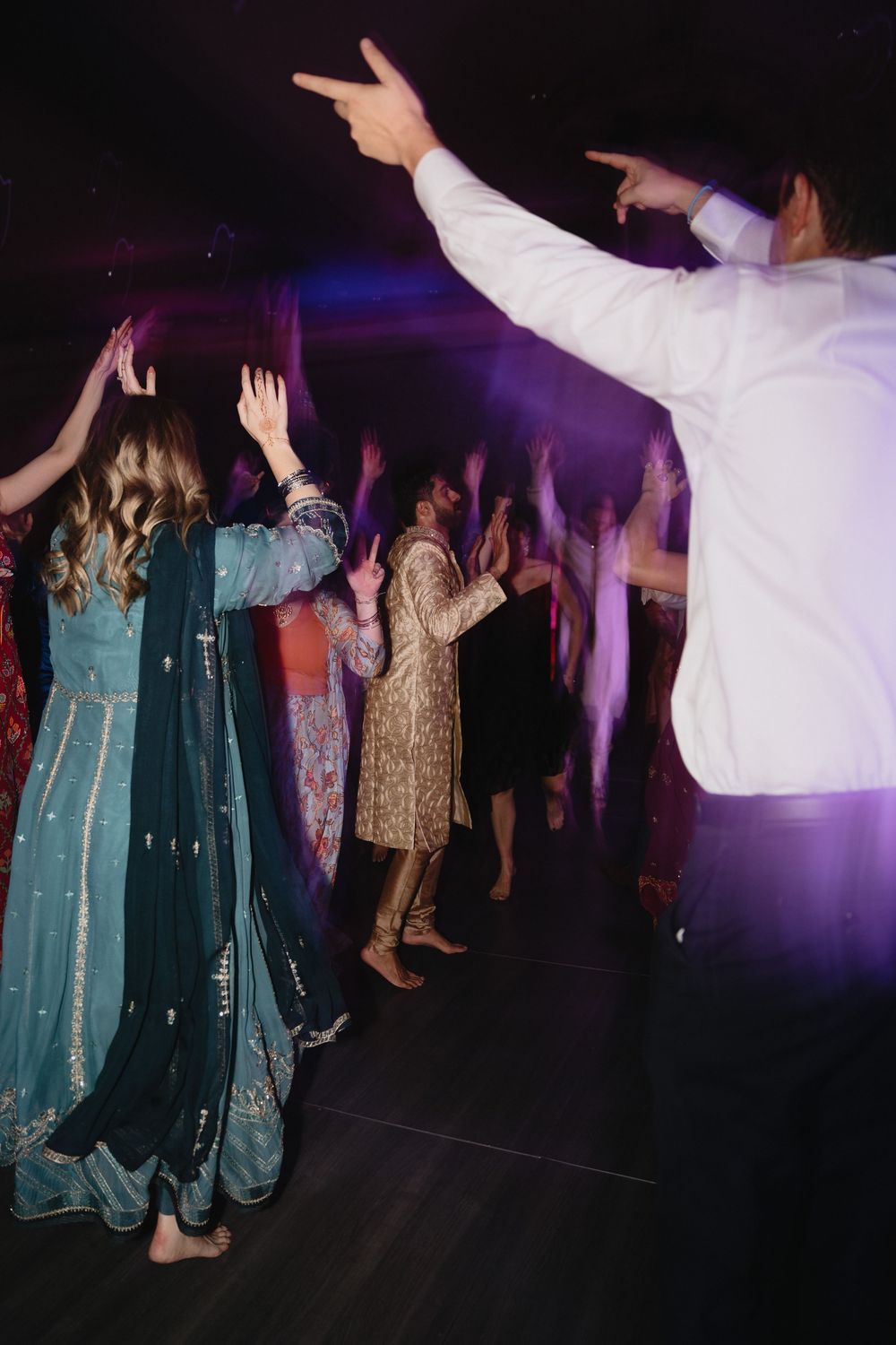 Wedding guests in traditional Indian attire dance with raised hands under purple party lights.