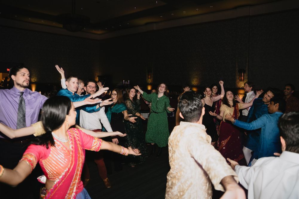 A large group of wedding guests in vibrant Indian attire enjoy a lively group dance at the reception.