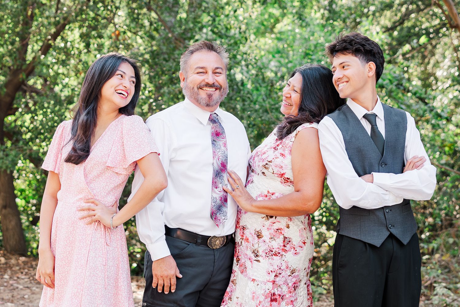 A group of wedding guests in floral dresses and formal attire laugh together in a lush outdoor garden setting.