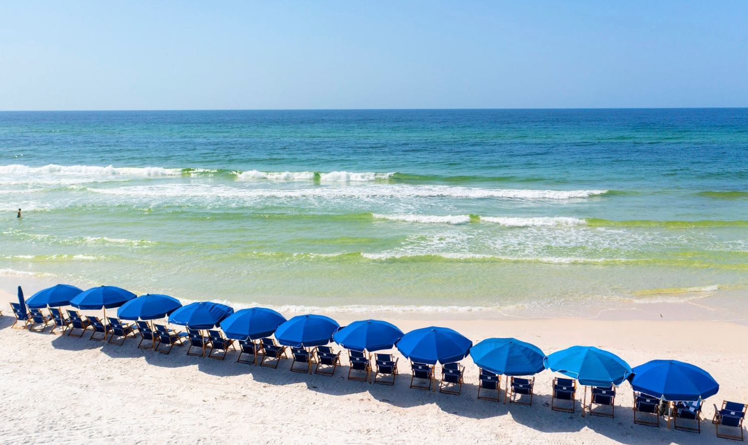 A row of blue beach umbrellas line a pristine white sand beach with turquoise ocean waves in the background.