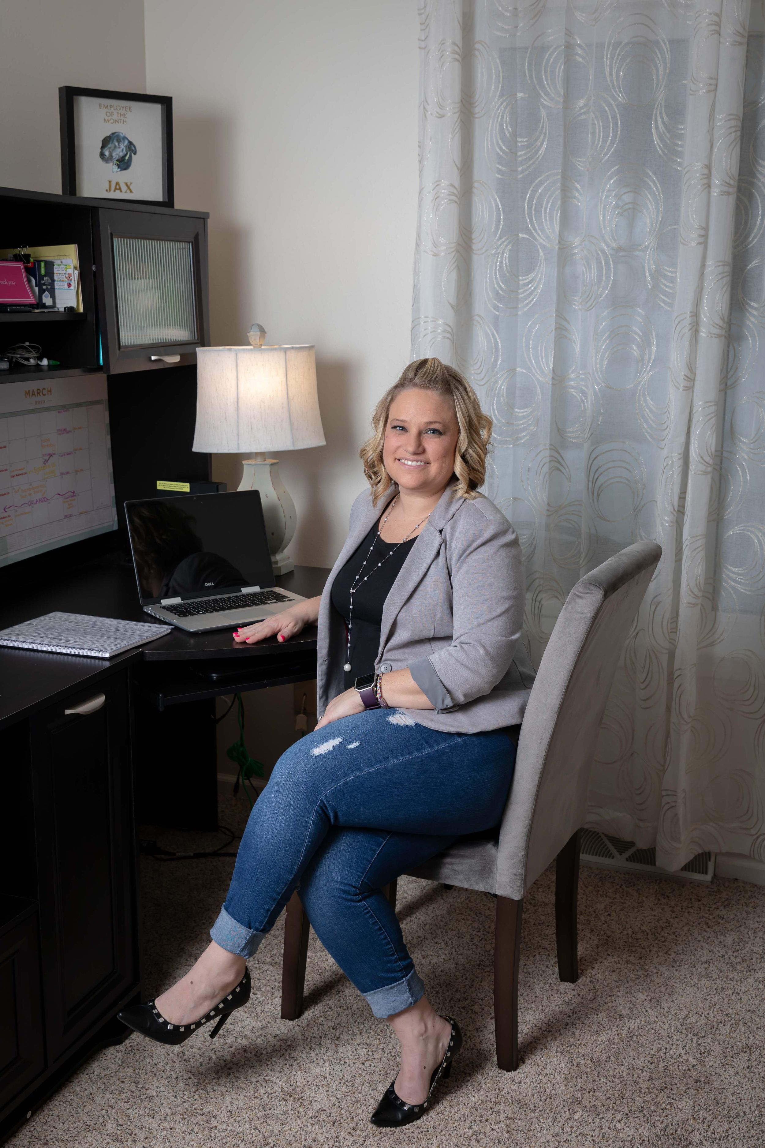 Professional working at home office desk wearing gray blazer and jeans in comfortable workspace setting.