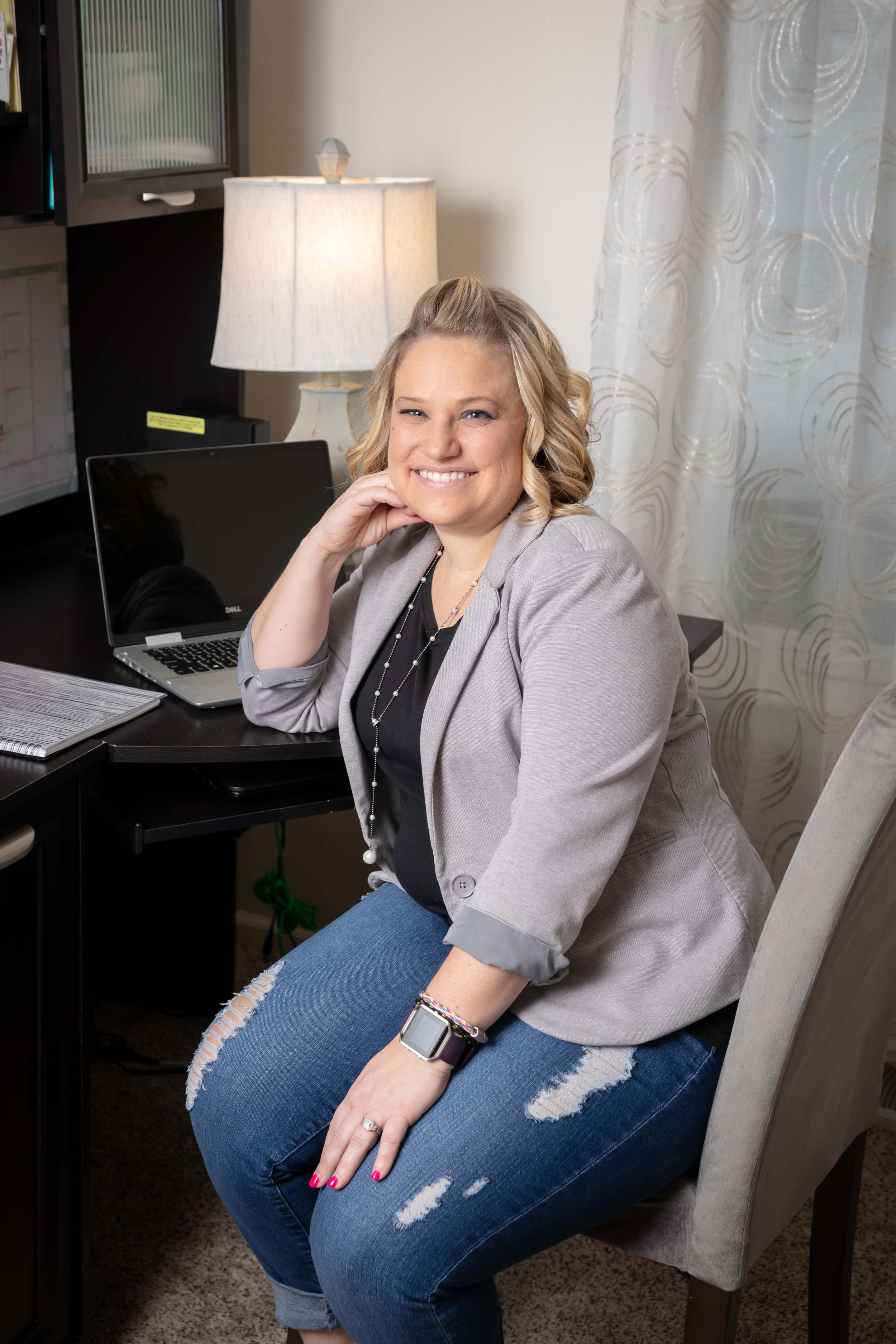 Person in ripped jeans and gray blazer poses casually on white couch in home office setting.