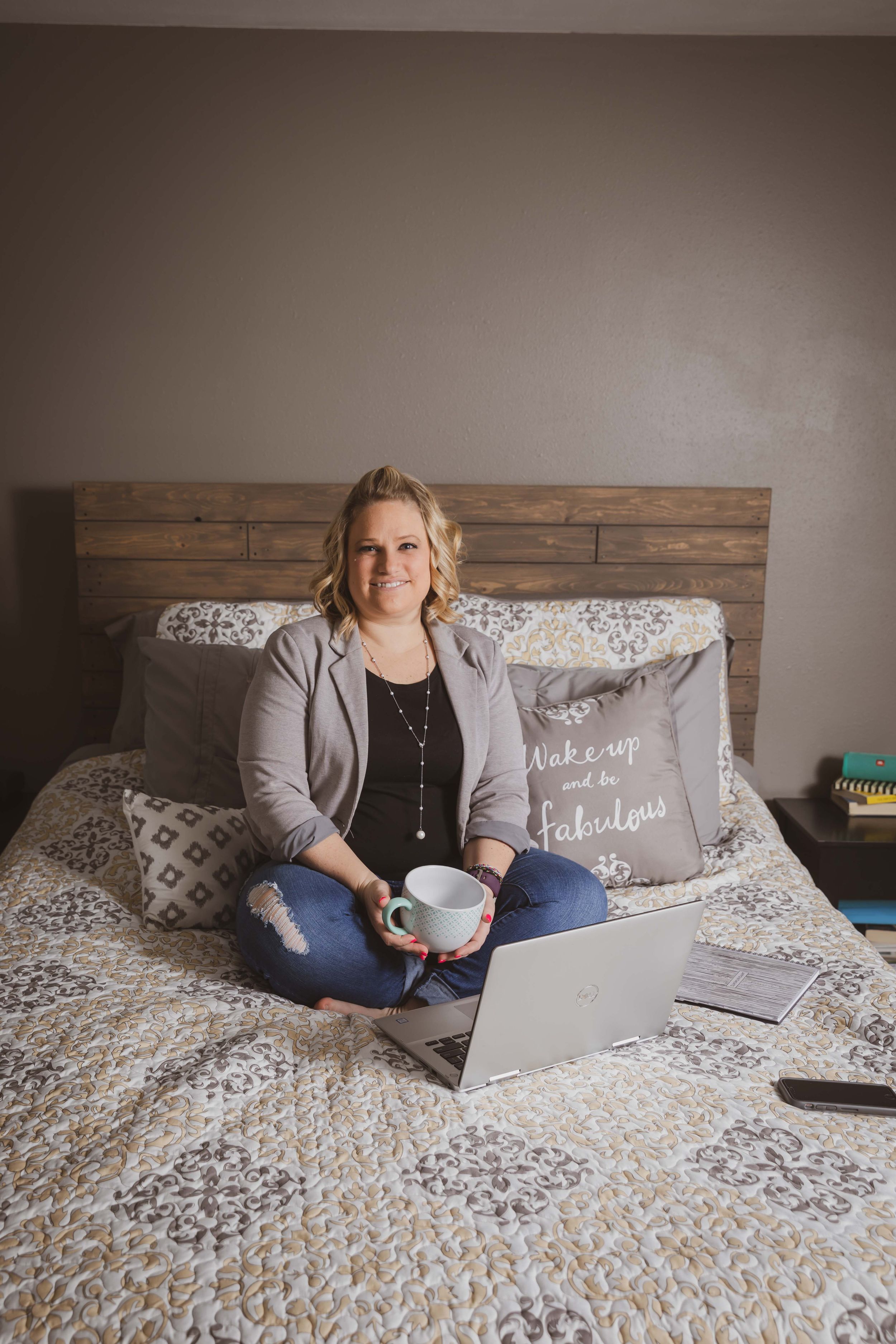 Person working on laptop while sitting cross-legged on a bed with decorative pillows and wooden headboard.