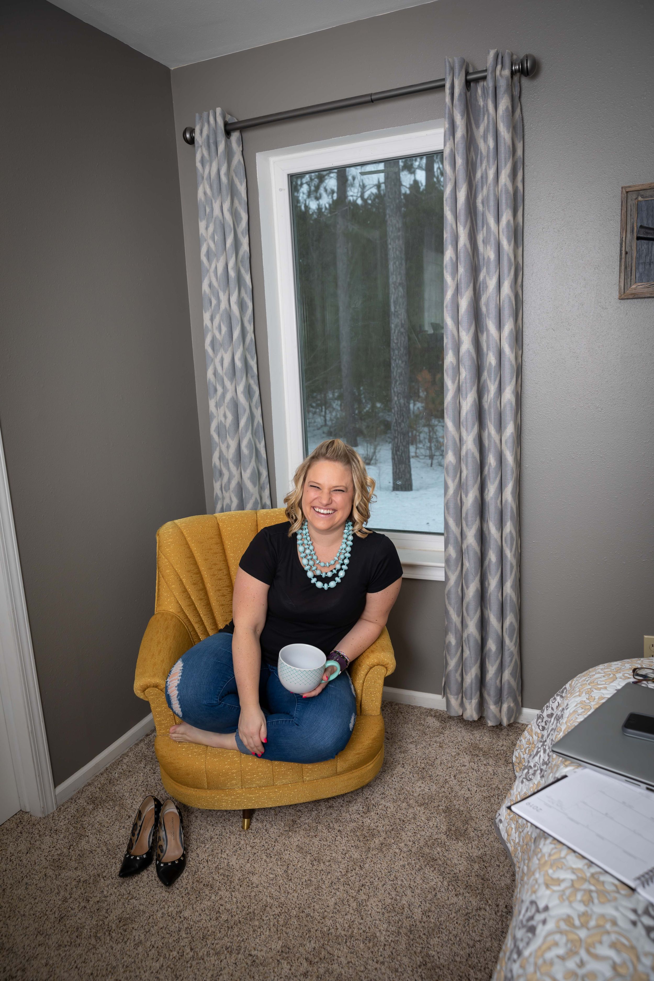 A comfortable yellow accent chair is positioned near a window with white curtains and gray walls.