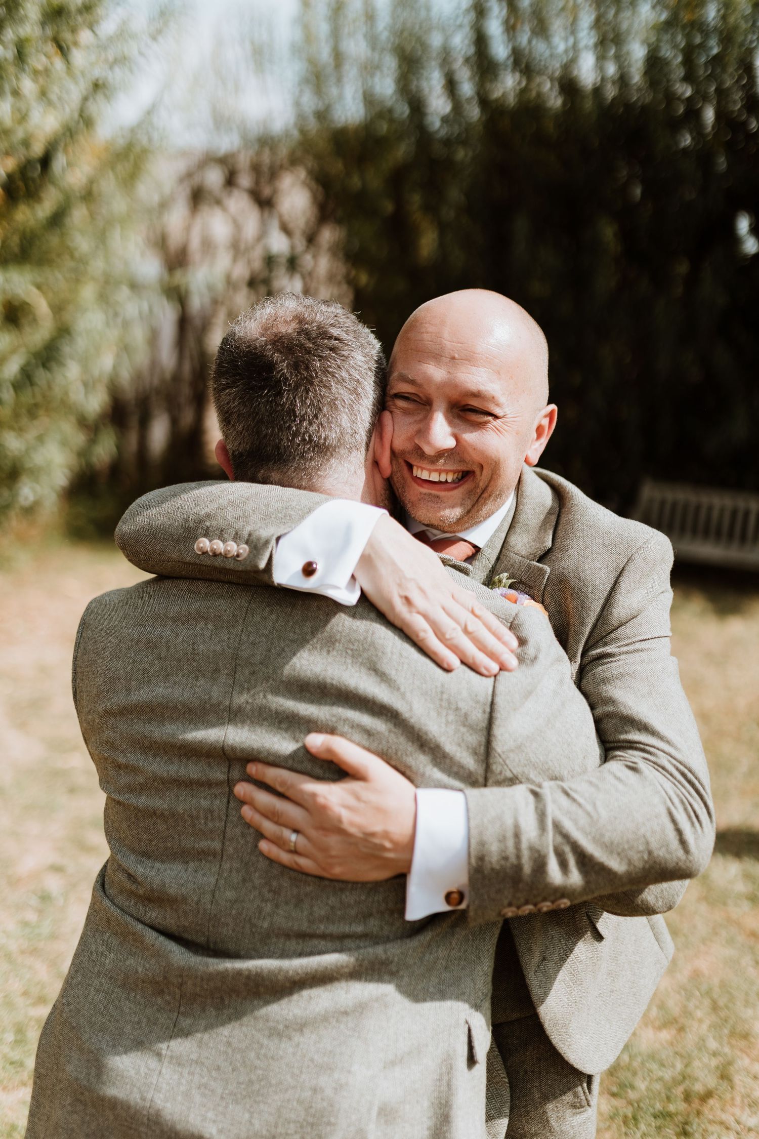 Two people in gray suits share an emotional embrace during what appears to be a wedding celebration outdoors.