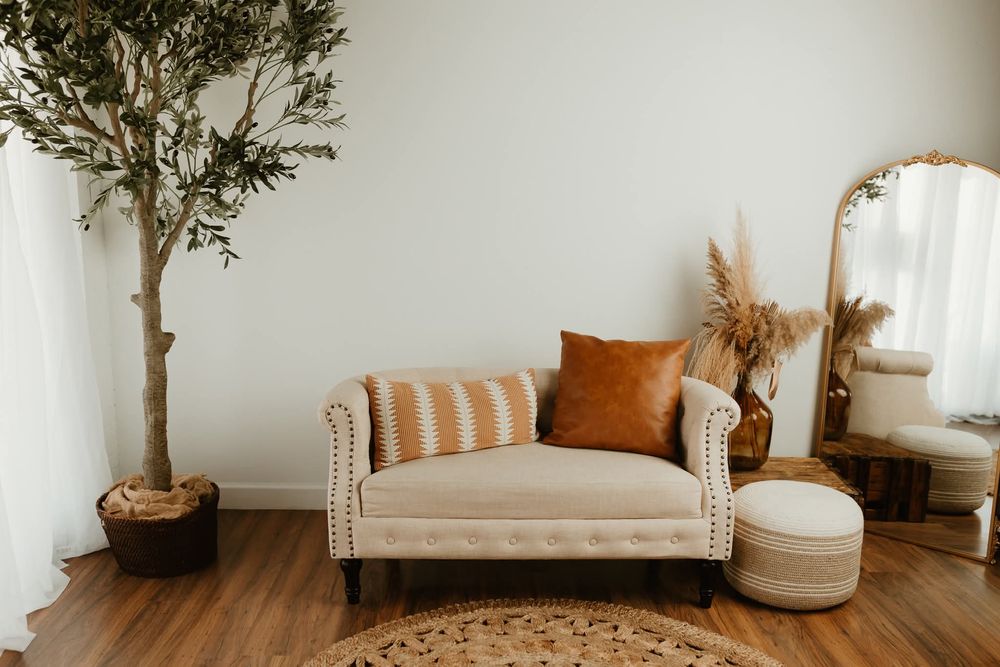 Boho living room with cream tufted loveseat, burnt orange pillow, pampas grass, gold mirror, and olive tree on wood floors.