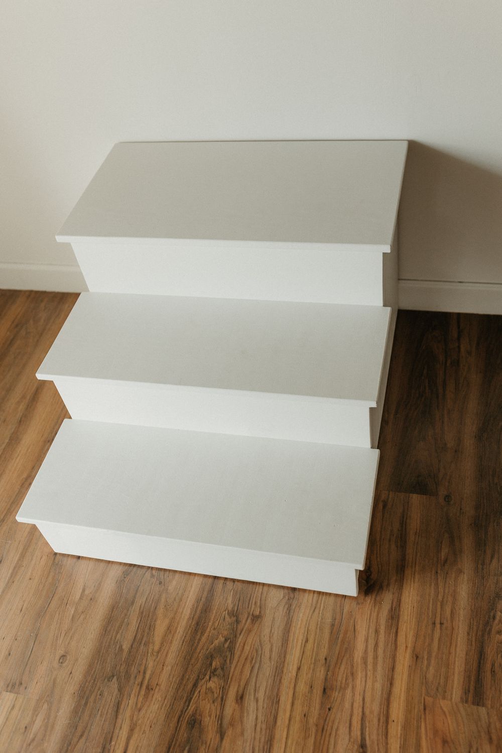 White three-tier pet stairs with a modern design, placed on a wooden floor against a white wall.