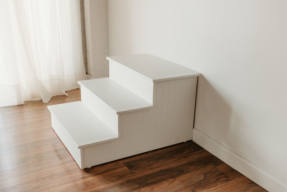 White wooden pet stairs with three steps and storage drawers, placed in a bright room with hardwood floors.