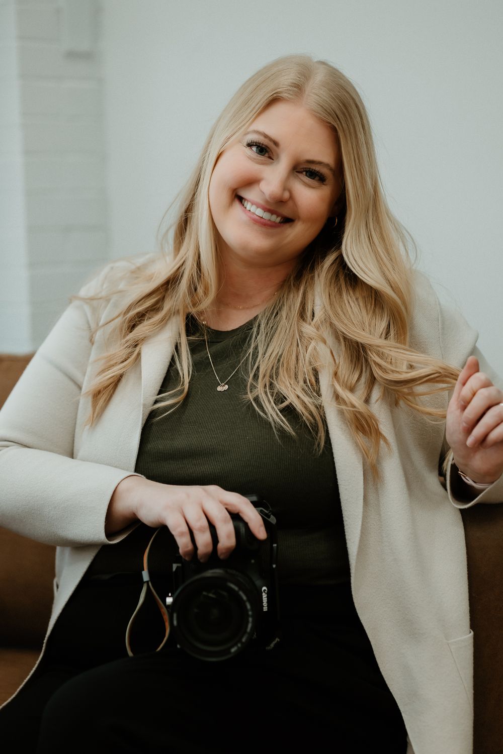 A photographer in a cream cardigan and black top poses with a camera in a bright indoor setting.
