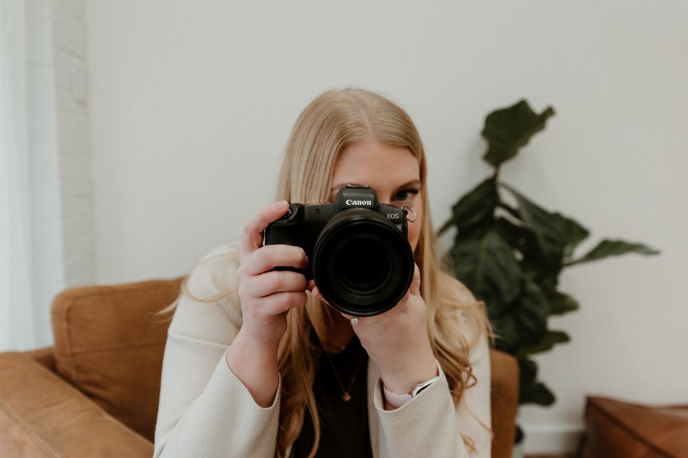 A person holds a black camera in front of a white wall with a green plant visible in the background.