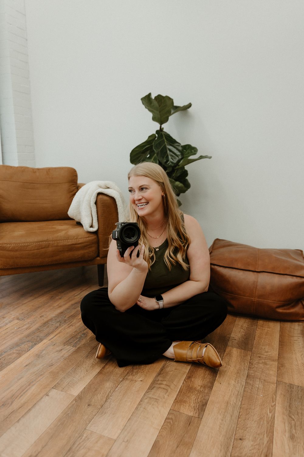 A photographer in a black dress sits on hardwood floors holding a camera in a bright minimalist studio setting.