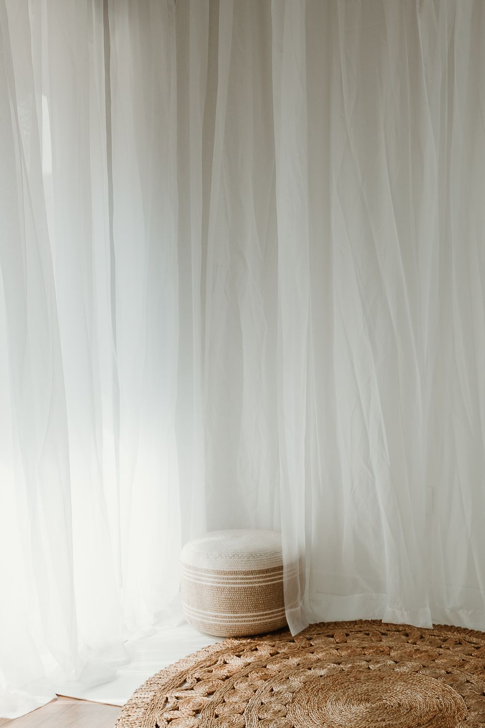 Sheer white curtains draped in a bright room with a woven rattan pouf and jute rug on hardwood floor.