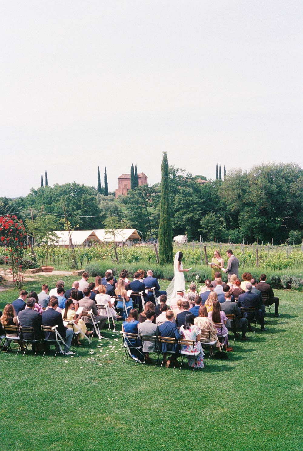 Outdoor wedding ceremony on lush green lawn with guests seated in rows, vineyard and rustic buildings in background.