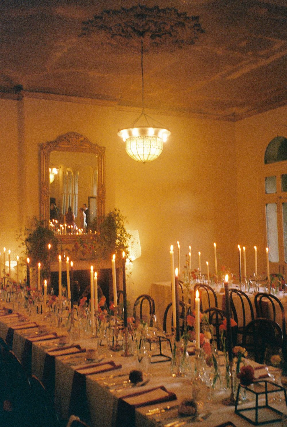 Elegant candlelit dining room with long tables, tall tapers, chandelier, and ornate mirror for a formal dinner event.