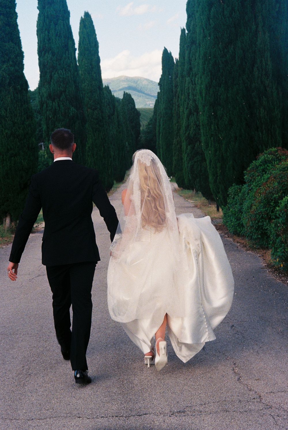 Bride and groom walking hand-in-hand down a cypress tree-lined path in Tuscany, Italy.